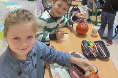 Chłopiec i dziewczynka siedzą w ławce w sali klasowej i prezentują swoje ludziki wykonane z dyni
