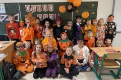 Zdjęcie grupowe uczniów klasy 1b ubranych w pomarańczowe kostiumy w rysunkami dyni. W tle tablica szkolna z napisem dzień dyni