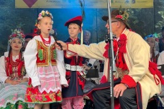 Uczniowie  stoją na scenie podczas śpiewania piosenki w towarzystwie siedzącego na krześle reżysera przedstawienia Józefa Plewniaka, który trzyma mikrofon przy ustach dziewczynki. W tle  sceny widać trzy kobiety w krakowskich strojach ludowych.