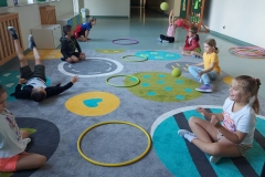 Zabawy z piłka i hula-hop. Uczniowie siedzą na dywanie, rzucają piłkę do obręczy.