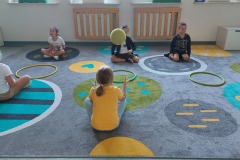 Zabawy z piłka i hula-hop. Uczniowie siedzą na dywanie, rzucają piłkę do obręczy.