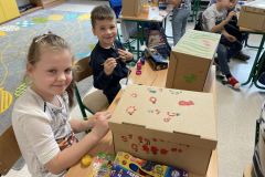 Chłopiec i dziewczynka siedzą w ławce, malują farbami kartonowe pudełka. Na pudełkach malowane są kwiaty i inne motywy roślinne