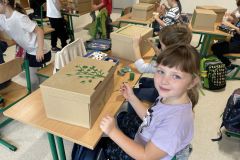 Dziewczynka siedzi w sali klasowej przy ławce i maluje kartonowe pudełko farbami