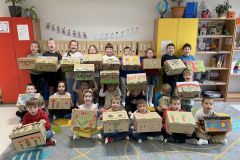 Uczniowie kl. 1b siedzą na dywanie w klasie i trzymają przed sobą kartonowe pudełka pomalowane farbami w motywy roślinne.