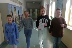 Trzy dziewczyny i chłopiec idą uśmiechnięci korytarzem szkolnym niosąc przygotowane ulotki dla młodszych uczniów.