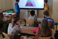 Uczniowie prezentują swoją pracę na tablicy multimedialnej. W tle widać innych uczniów słuchających swoich kolegów. Na ekranie widać „piramidę żywieniową”.