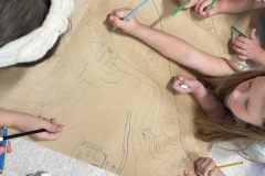Fragment szkicu otoczenia rzeki Niemen wykonywany przez kilkoro dzieci na wspólnym arkuszu szarego papieru. Szkic przedstawia dopływy rzeki, budynki, drzewa oraz ludzką postać.
