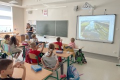 Uczniowie klasy 1b siedząc w ławkach  sali lekcyjnej i oglądają prezentację pt. Nad Niemnem  wyświetlaną na tablicy interaktywnej przez bibliotekarkę. Wyświetlone jest zdjęcie rzeki Niemen  wraz z jej otoczeniem.