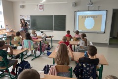 Uczniowie klasy 1b siedząc w ławkach  sali lekcyjnej i oglądają prezentację pt. Nad Niemnem  wyświetlaną na tablicy interaktywnej przez bibliotekarkę.